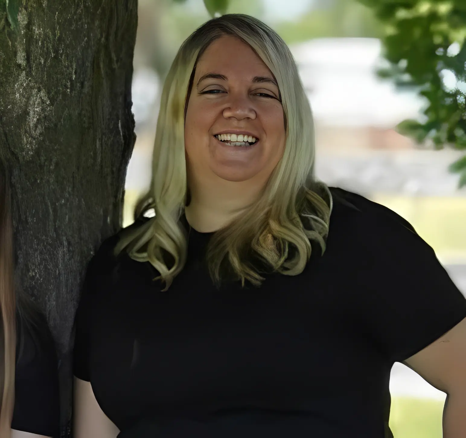 A smiling woman with long blonde hair standing by a tree.